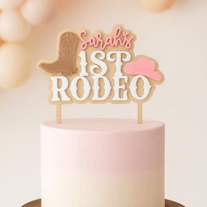 May include: A cake topper featuring the text Sarah's 1ST RODEO in white and pink, accompanied by a brown cowboy boot and a pink cowboy hat icon. The topper is mounted on two thin sticks and is positioned atop a two-toned light pink and white cake.