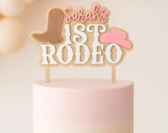 May include: A cake topper featuring the text Sarah's 1ST RODEO in white and pink, accompanied by a brown cowboy boot and a pink cowboy hat icon. The topper is mounted on two thin sticks and is positioned atop a two-toned light pink and white cake.