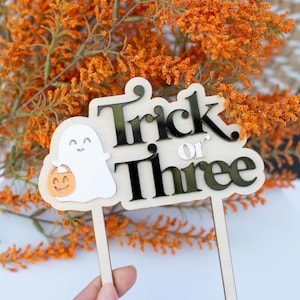 May include: A wooden cake topper featuring the text Trick or Three in black lettering, accompanied by a white ghost figure holding a small orange pumpkin bucket. The design is mounted on two wooden stakes for insertion into a cake or other surface.