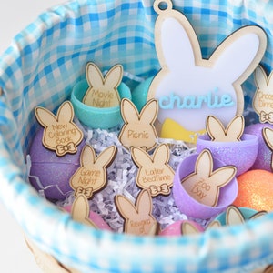 May include: A blue and white gingham-lined basket filled with Easter-themed decorations. The basket contains wooden bunny-shaped tags with activity labels like "Movie Night" and "New Coloring Book." There are also decorative eggs in various colors.