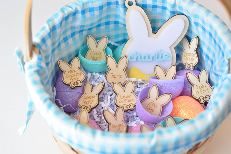 May include: A blue and white gingham-lined basket filled with Easter-themed decorations. The basket contains wooden bunny-shaped tags with activity labels like "Movie Night" and "New Coloring Book." There are also decorative eggs in various colors.