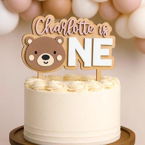 May include: A white frosted cake with a wooden cake topper that reads "Charlotte is ONE" with a bear design. The cake sits on a wooden cake stand, with a backdrop of white, brown, and pink balloons.