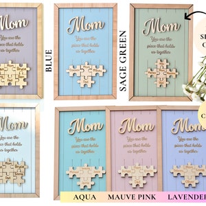 Puede incluir: Arte de pared enmarcado en madera con la palabra "Mom" en cursiva, la frase "You are the piece that holds us together" y un rompecabezas de madera con nombres. Disponible en ciruela, azul, verde salvia, blanco, aguamarina, rosa malva y lavanda.