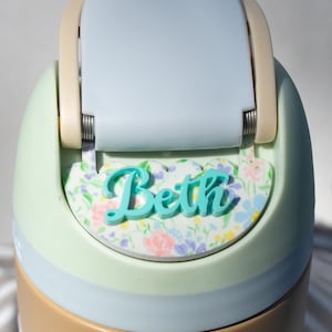 Puede incluir: Una botella de agua personalizada con tapa azul claro y cuerpo verde menta. El nombre "Beth" se muestra en escritura turquesa sobre una base con estampado floral. La botella tiene una banda de color beige en la base.