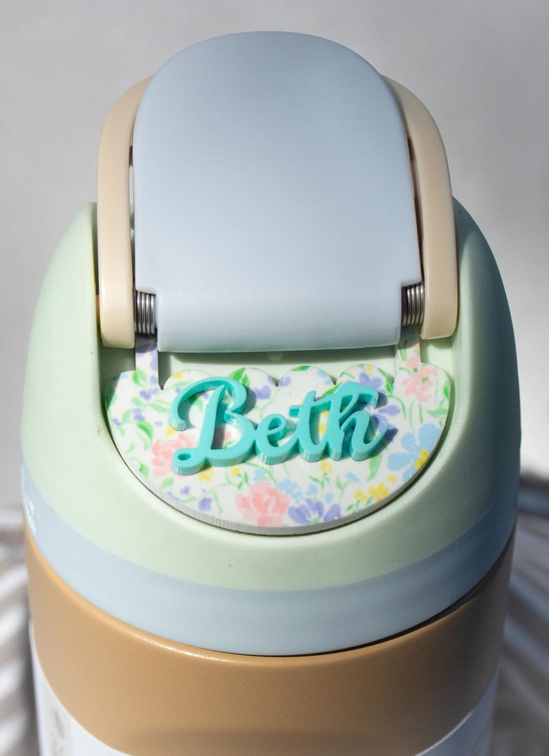 Puede incluir: Una botella de agua personalizada con tapa azul claro y cuerpo verde menta. El nombre "Beth" se muestra en escritura turquesa sobre una base con estampado floral. La botella tiene una banda de color beige en la base.