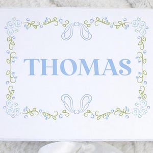 May include: White box with the name "THOMAS" in light blue, framed by a floral and ribbon design in light blue and green. The box is likely for a gift or keepsake.