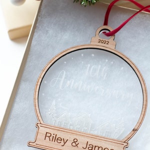 May include: A wooden Christmas ornament in the shape of a snow globe, with the text "10th Anniversary" and "Riley & James". The ornament is in a gift box with red ribbon and decorative berries.
