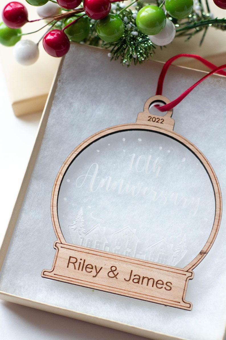 May include: A wooden Christmas ornament in the shape of a snow globe, with the text "10th Anniversary" and "Riley & James". The ornament is in a gift box with red ribbon and decorative berries.