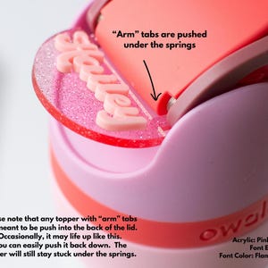 Puede incluir: Primer plano de una tapa de botella de agua rosa y roja con una cubierta acr&iacute;lica personalizada. La cubierta, que deletrea "Halley" en letras rosas, est&aacute; unida a la tapa. La tapa es rosa y roja, con el texto "Arm tabs are pushed under the springs."