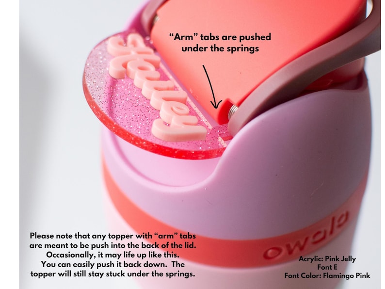 Puede incluir: Primer plano de una tapa de botella de agua rosa y roja con una cubierta acr&iacute;lica personalizada. La cubierta, que deletrea "Halley" en letras rosas, est&aacute; unida a la tapa. La tapa es rosa y roja, con el texto "Arm tabs are pushed under the springs."