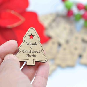 K&ouml;nnte beinhalten: Ein h&ouml;lzerner Weihnachtsbaum-Anh&auml;nger mit dem Text "Watch a Christmas Movie" in brauner Schrift. Der Anh&auml;nger wird in einer Hand vor einem verschwommenen Hintergrund gehalten.