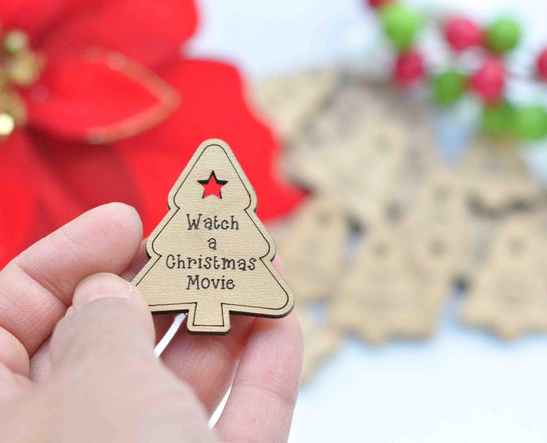K&ouml;nnte beinhalten: Ein h&ouml;lzerner Weihnachtsbaum-Anh&auml;nger mit dem Text "Watch a Christmas Movie" in brauner Schrift. Der Anh&auml;nger wird in einer Hand vor einem verschwommenen Hintergrund gehalten.