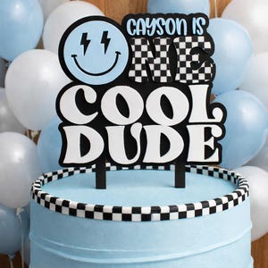 Könnte beinhalten: Ein blauer Kuchen mit einem schwarz-weißen karierten Rand, gekrönt von einem Tortenaufsatz mit der Aufschrift "Cayson is ONE COOL DUDE". Der Aufsatz zeigt ein Smiley-Gesicht mit Blitzen und die karierte Zahl eins.
