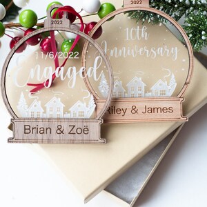 May include: Two wooden Christmas ornaments with clear centers and a town silhouette design. One says "Engaged" with a date, the other says "10th Anniversary". Both have names and the year 2022.