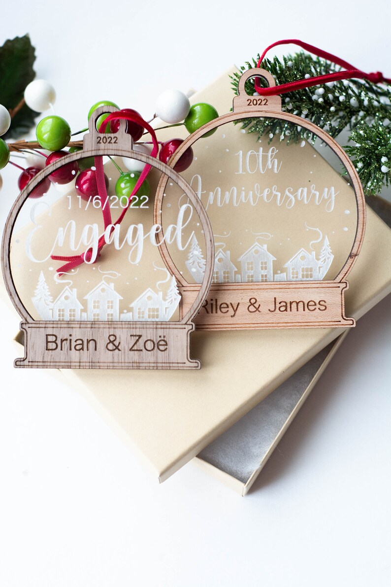 May include: Two wooden Christmas ornaments with clear centers and a town silhouette design. One says "Engaged" with a date, the other says "10th Anniversary". Both have names and the year 2022.