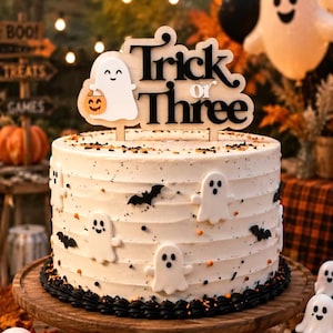 May include: A round white frosted cake decorated with small white ghost and black bat cutouts, topped with a wooden cake topper that reads Trick or Three and features a small ghost holding a pumpkin bucket, set on a wooden stand.