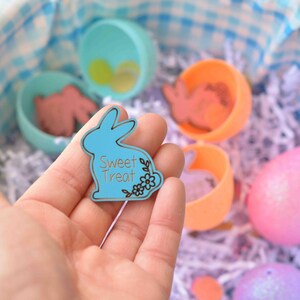 May include: A light blue bunny-shaped Sweet Treat tag with floral accents is held over a basket filled with Easter eggs and treats. The basket has a blue and white checkered lining. Other Easter eggs are visible.