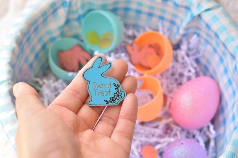 May include: A light blue bunny-shaped Sweet Treat tag with floral accents is held over a basket filled with Easter eggs and treats. The basket has a blue and white checkered lining. Other Easter eggs are visible.