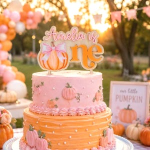 May include: A two-tiered cake topper made of light wood featuring the text Amelia is one in a script and block font. The design includes a large orange pumpkin illustration with a pink bow. The topper is positioned on a pink and orange cake.
