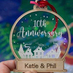May include: Wooden 10th Anniversary ornament with a clear center, featuring a winter village scene. The ornament has the text "10th Anniversary" and "Katie & Phil" with the year "2022" on top. A red ribbon is attached.