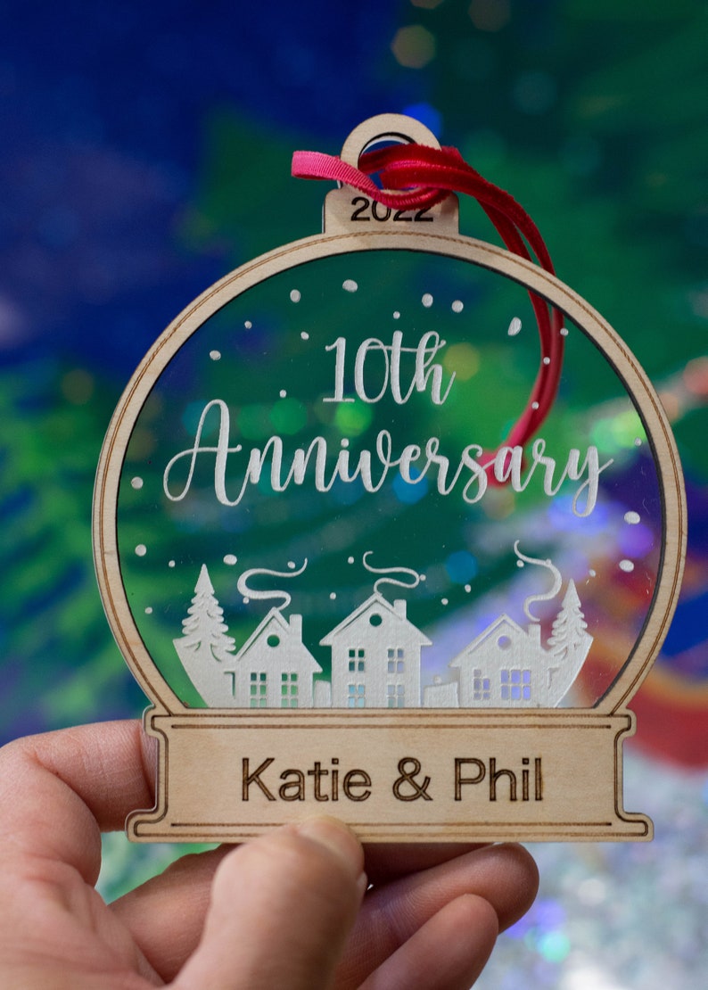 May include: Wooden 10th Anniversary ornament with a clear center, featuring a winter village scene. The ornament has the text "10th Anniversary" and "Katie & Phil" with the year "2022" on top. A red ribbon is attached.