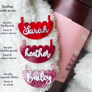 Puede incluir: Tres etiquetas de nombre personalizadas de acr&iacute;lico en forma de vieira en rojo, burdeos y purpurina burdeos. Los nombres "Sarah", "heather" y "Bailey" se muestran en escritura blanca. Las etiquetas est&aacute;n sobre una superficie blanca y esponjosa.