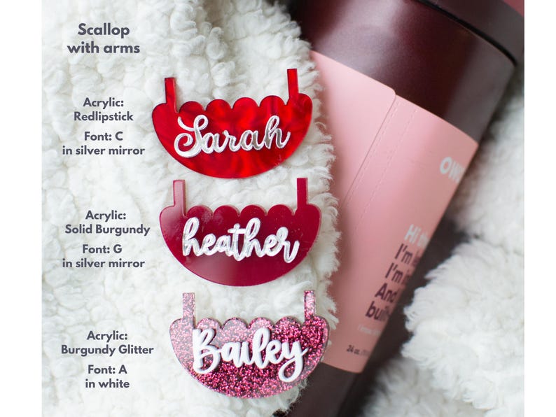 Puede incluir: Tres etiquetas de nombre personalizadas de acr&iacute;lico en forma de vieira en rojo, burdeos y purpurina burdeos. Los nombres "Sarah", "heather" y "Bailey" se muestran en escritura blanca. Las etiquetas est&aacute;n sobre una superficie blanca y esponjosa.