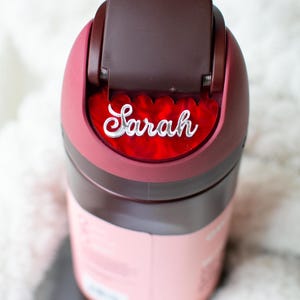 Puede incluir: Una botella de agua aislada de color granate y rosa con el nombre "Sarah" en escritura blanca. La botella tiene una tapa abatible y un cuerpo rosa. El nombre "Sarah" se muestra en un acento rojo en forma de coraz&oacute;n.