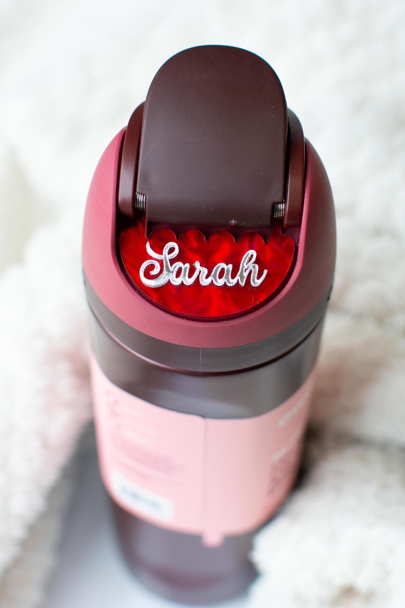 Puede incluir: Una botella de agua aislada de color granate y rosa con el nombre "Sarah" en escritura blanca. La botella tiene una tapa abatible y un cuerpo rosa. El nombre "Sarah" se muestra en un acento rojo en forma de coraz&oacute;n.