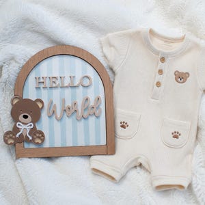 May include: A cream-coloured waffle-knit romper with three wooden buttons, a small bear embroidery, and two pockets with paw prints, alongside an arched wooden sign featuring blue stripes, a bear cutout, and the text HELLO World.
