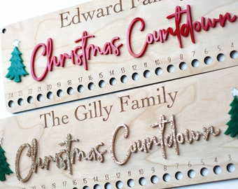 May include: Two rectangular light wood plaques featuring the engraved text Edward Family and The Gilly Family. Each plaque has a 3D Christmas Countdown sign, one in red acrylic and one in gold glitter, with green tree icons and numbered holes.
