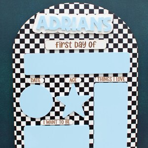 May include: An arched sign with a black and white checkered pattern featuring the name ADRIANS in light blue letters. It includes sections labeled First Day Of, Date, Age, Things I Love, and I Want To Be, with a dry erase marker and a Last Day Of sign.