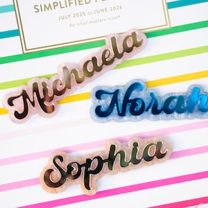 May include: Three name tags with a script font are displayed on a striped background. The names are "Michaela", "Nora", and "Sophia". The "Michaela" tag is pink with gold lettering, "Nora" is blue with a silver outline, and "Sophia" is tan with gold lettering.