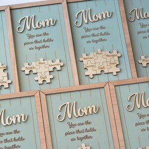 Puede incluir: Arte de pared enmarcado en madera con la palabra "Mom" en fuente cursiva y la frase "You are the piece that holds us together." Cada pieza tiene un dise&ntilde;o de rompecabezas con nombres. El fondo es azul claro.