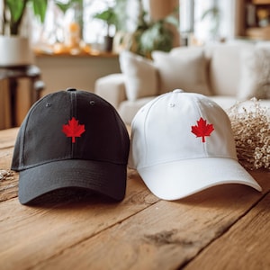 May include: A pair of baseball caps, one black and one white, both featuring a red maple leaf design. The caps are presented on a wooden table, suitable for casual wear.