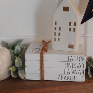 May include: A stack of four white books with black lettering. The top book says "BOOKYARD" and the other books say "TAYLOR", "LINDSAY", "HANNAH", and "CHARLOTTE". The books are tied together with twine. A white ceramic house and a black metal star are on the shelf behind the books.