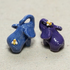 Puede incluir: Dos pequeñas figuritas de elefante de cerámica, una azul y otra morada, con un detalle de abeja amarilla en cada una.
