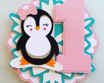 May include: A pink and white layered cake topper featuring a cartoon penguin with a pink bow, black and white body, and orange feet. A pink number one is next to the penguin. The topper is surrounded by teal and white accents.