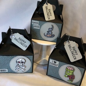 May include: Three black Halloween gift boxes with grey detailing. Each box has a different Halloween graphic: a skull and crossbones, a witch's hat, and a Frankenstein's monster. Each box has a tag that says "Happy Halloween".