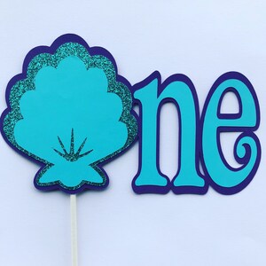 May include: A turquoise blue seashell with glitter accents and the word "one" in turquoise blue with a purple outline. The seashell is on a stick.
