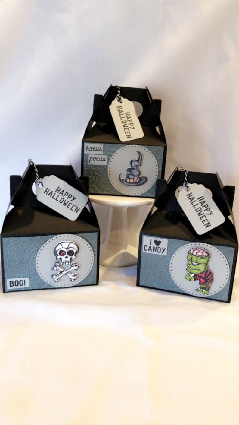 May include: Three black Halloween gift boxes with tags. Each box has a different design: a witch's hat, a skull and crossbones, and a cartoon zombie. The tags say "Happy Halloween".