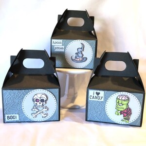 May include: Three black Halloween gift boxes with handles. Each box has a circular design with a different graphic: a witch's hat, a skull and crossbones, and Frankenstein's monster. Text includes "BOO!", "hocus pocus", and "I &hearts; CANDY".