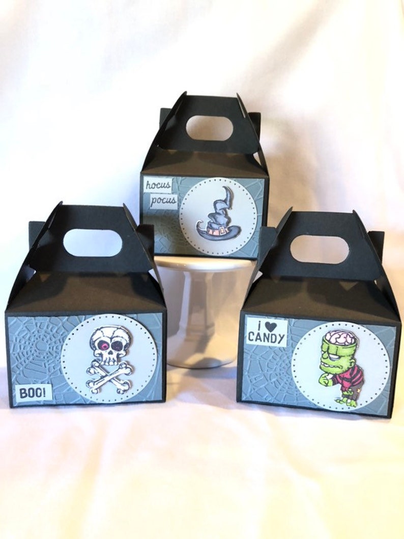 May include: Three black Halloween gift boxes with handles. Each box has a circular design with a different graphic: a witch's hat, a skull and crossbones, and Frankenstein's monster. Text includes "BOO!", "hocus pocus", and "I &hearts; CANDY".