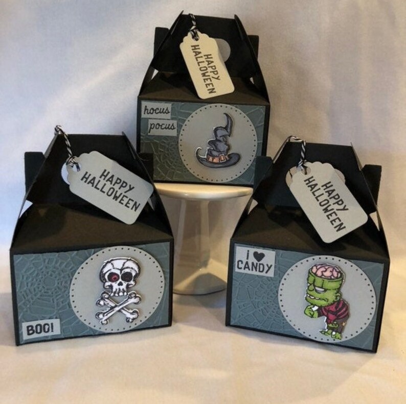 May include: Three black Halloween gift boxes with white tags saying "Happy Halloween." Each box has a different Halloween graphic: a witch's hat, a skull and crossbones, and a zombie. The boxes have a spiderweb design.