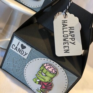 May include: A black and grey Halloween gift box with a tag saying "HAPPY HALLOWEEN". The box features a grey spiderweb design and a sticker that says "I ❤️ CANDY". A cartoon zombie is on the front.