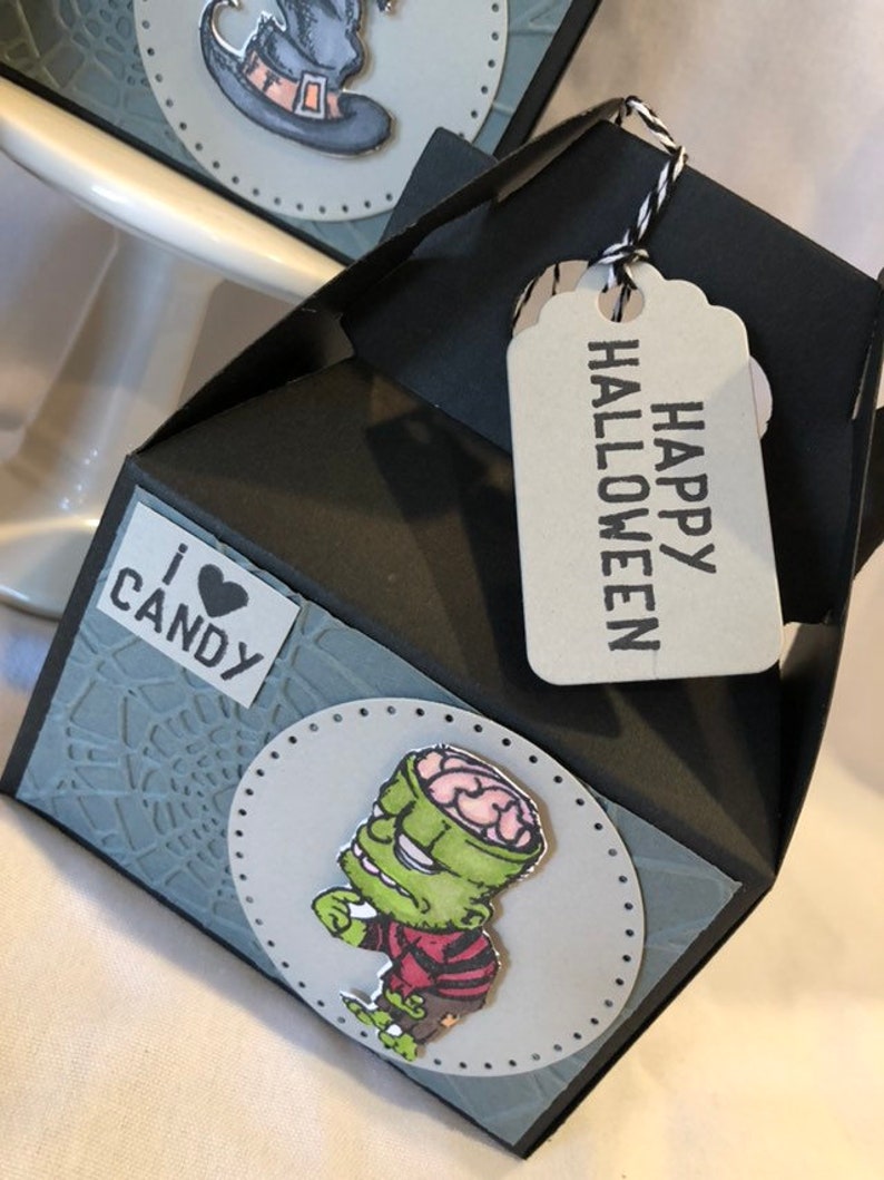 May include: A black and grey Halloween gift box with a tag saying "HAPPY HALLOWEEN". The box features a grey spiderweb design and a sticker that says "I ❤️ CANDY". A cartoon zombie is on the front.