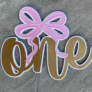 May include: Gold glitter "one" with a pink bow on top. The number one is outlined in white. This is a cake topper.