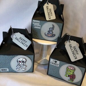 May include: Three black Halloween gift boxes with decorative oval stickers featuring a skull and crossbones, a witch's hat, and a zombie. Each box has a tag that says "Happy Halloween".
