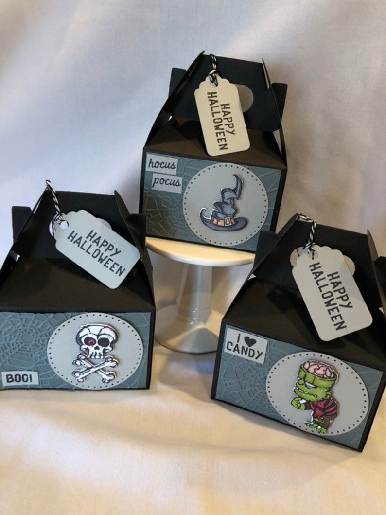 May include: Three black Halloween gift boxes with decorative oval stickers featuring a skull and crossbones, a witch's hat, and a zombie. Each box has a tag that says "Happy Halloween".