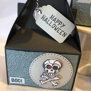 May include: A black Halloween gift box with a skull and crossbones design. The box has a grey tag that says "HAPPY HALLOWEEN". The box also has a grey spiderweb design and the word "BOO!"